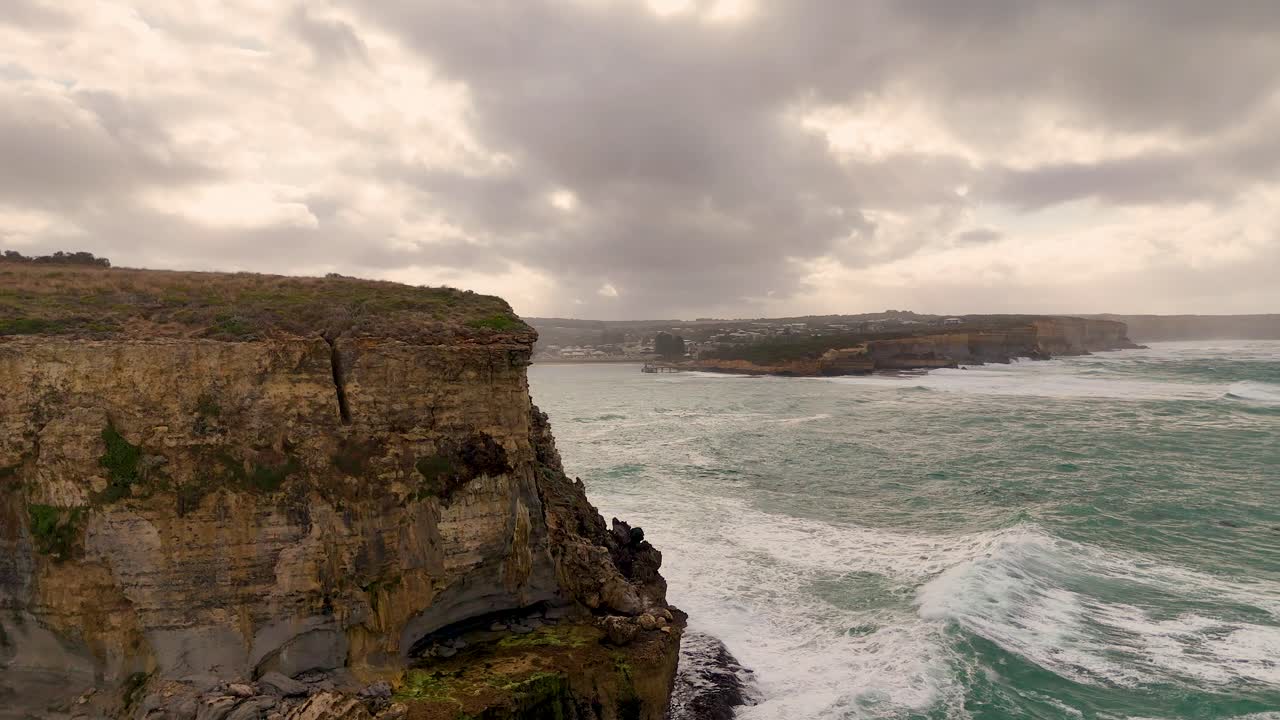Drone footage captures dramatic cliffs and turbulent ocean waves under cloudy skies at Port Campbell, Australia