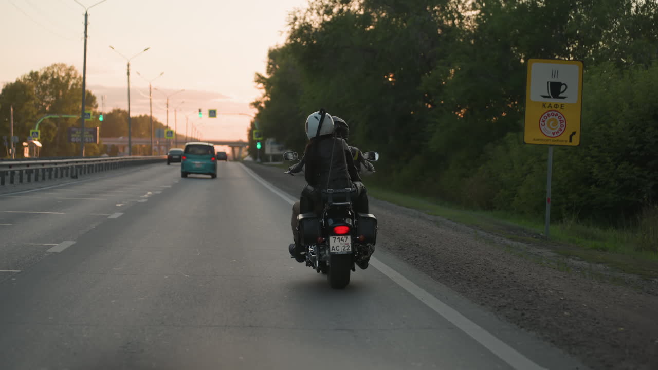 Motorcycle rider and passenger wearing helmets travel along highway at sunset, moving past roadside cafe sign while surrounded by cars, illuminated by warm golden light highlighting evening journey