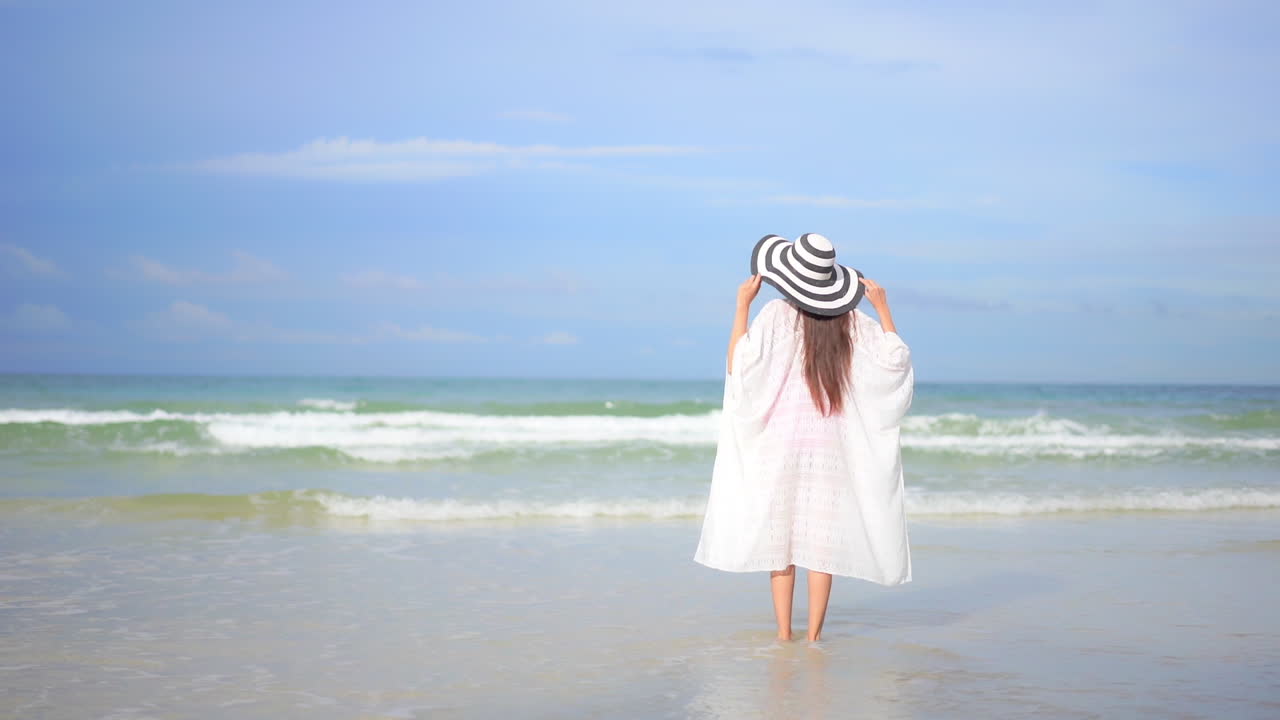 mujer asiática caminando en agua de mar poco profunda cuando grandes mareas espumosas ruedan en la playa de arena en verano, usando un vestido blanco y un sombrero de rayas, tocando el sombrero con ambas manos - vista trasera en cámara lenta