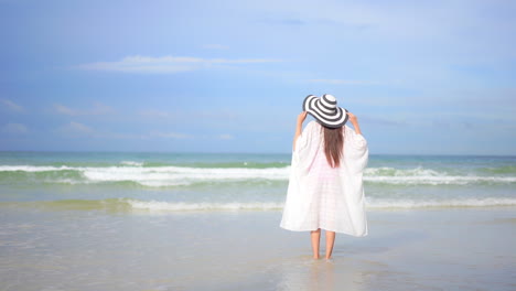 Asian-woman-walking-in-shallow-seawater-when-big-foamy-tides-roll-on-sandy-beach-in-summer,-wearing-a-white-sundress-and-stripy-sunhat,-touching-hat-with-both-hands---slow-motion-back-view