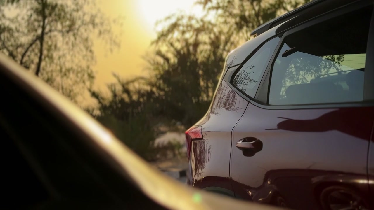 Scatto di un SUV scuro parcheggiato durante un bellissimo tramonto all'ora d'oro. La calda luce del sole filtra attraverso gli alberi sullo sfondo, riflettendosi sulla carrozzeria lucida del veicolo in un parcheggio all'aperto.