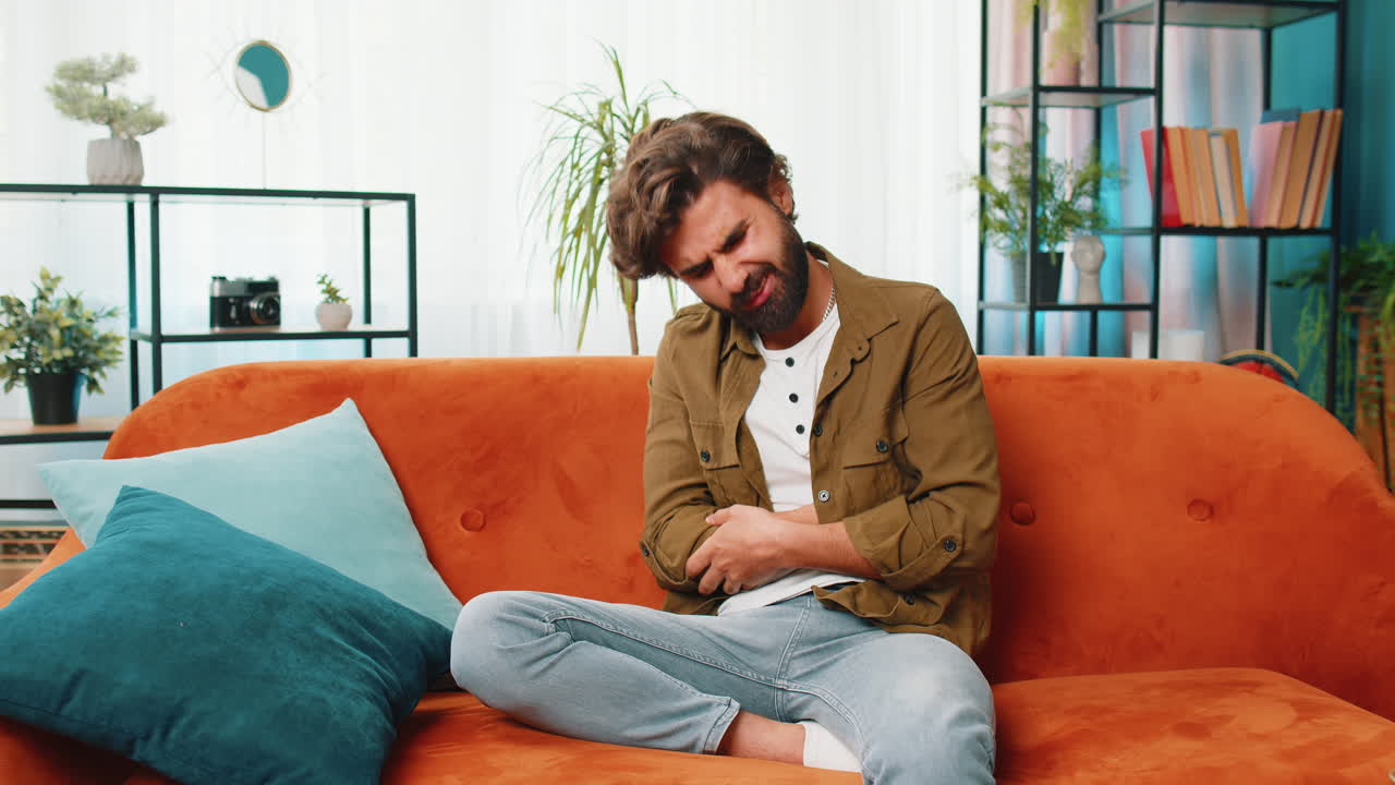 Arabian man sits on sofa feeling sudden strong abdominal stomach ache, gastritis problem, diarrhea
