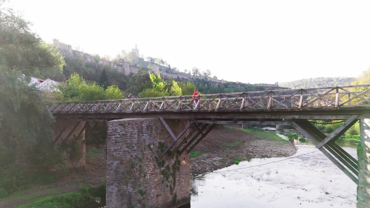 Man walking on Vladishki Most with Tsarevets fortress in background, zoom shot capturing medieval Veliko Tarnovo charm.