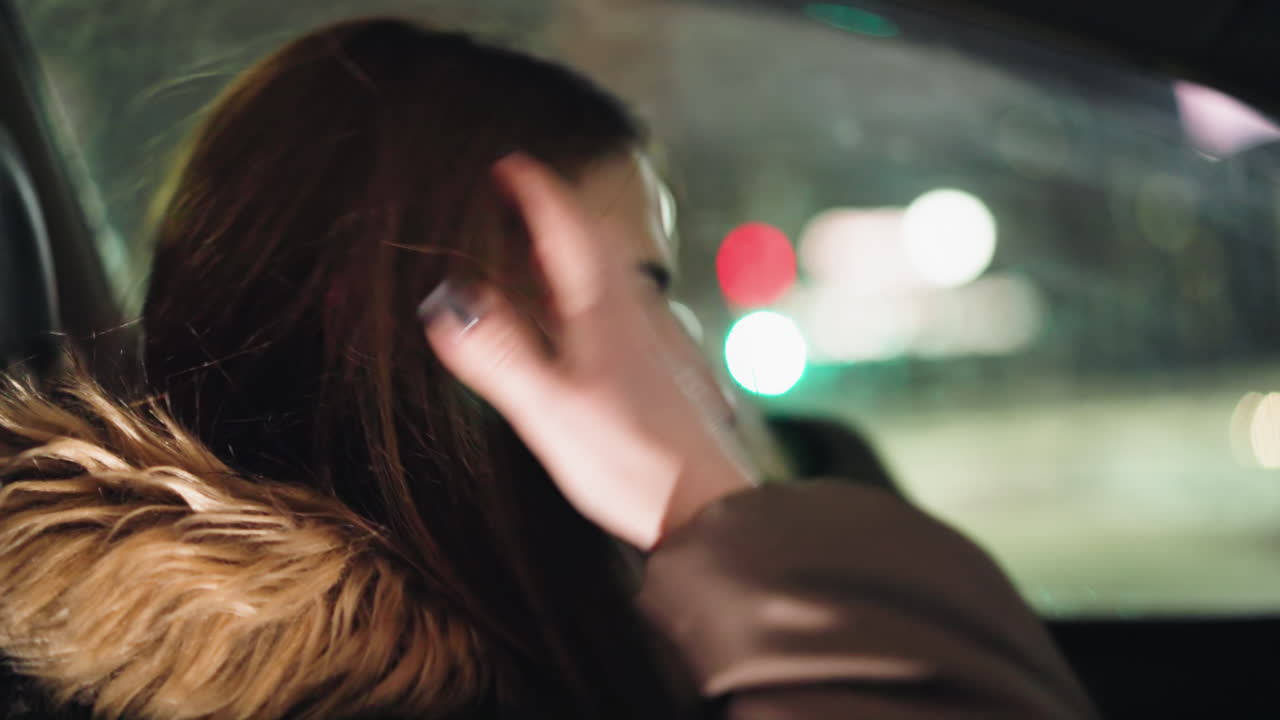 First-person view of a woman in a winter coat with a fur hood, inside a car. She has taken off her head warmer and is shaking her head. The background shows blurry city lights