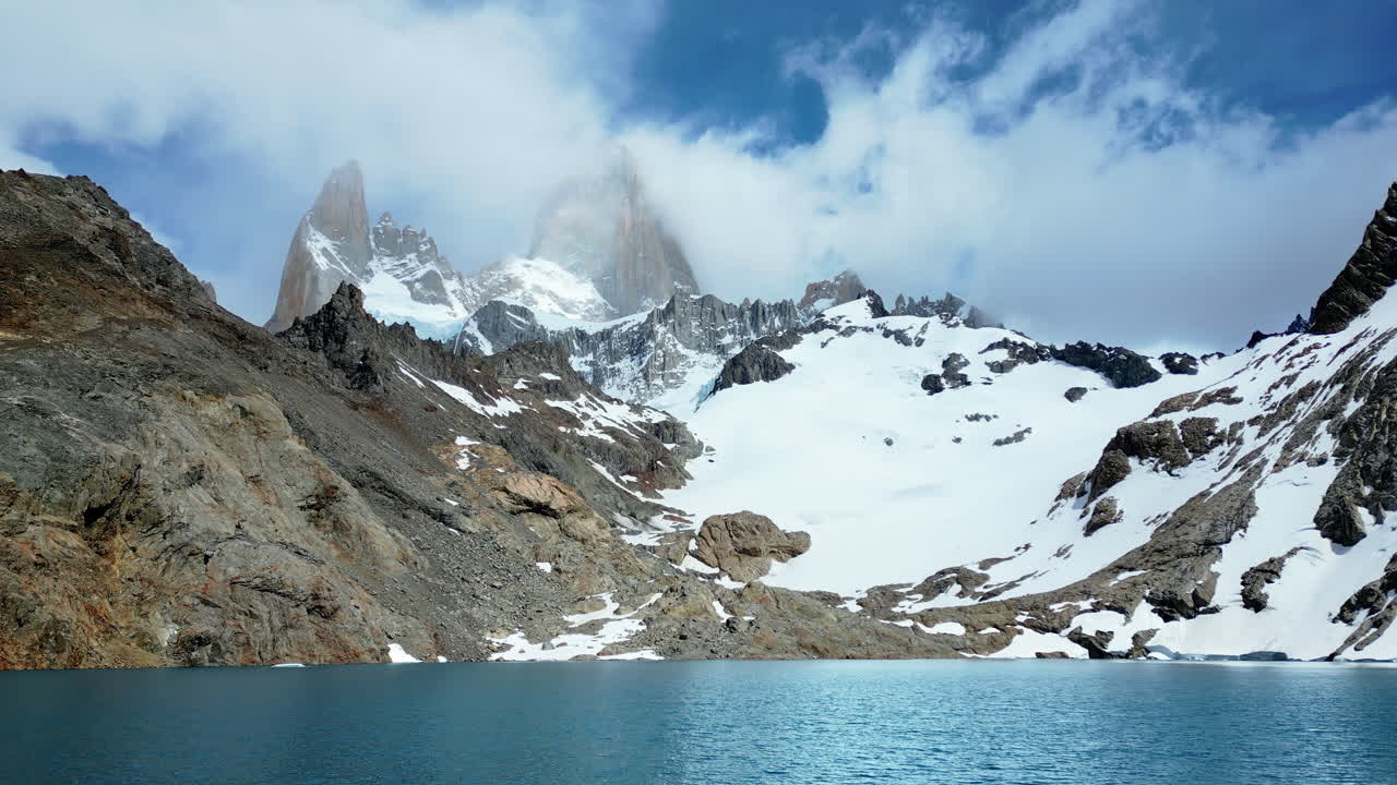 Aerial drone view of a turquoise mountain lake surrounded by rugged terrain and snowfields beneath the Fitz Roy massif
