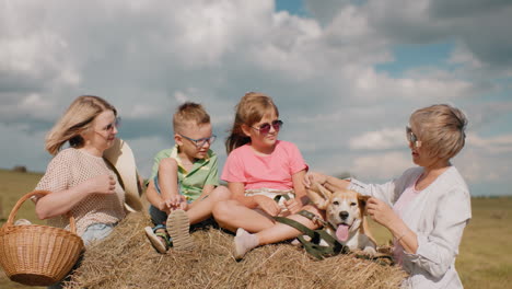 la familia sonriente disfruta del campo mientras la abuela estira juguetonamente las orejas del perro, los niños se relajan en el pajar, la mujer con la canasta se ríe, el campo soleado con balas de heno y el cielo azul
