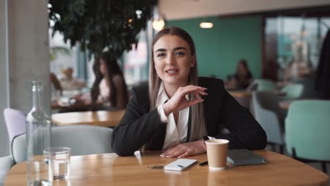 Schöne-Junge-Frau-Sitzt-In-Einem-Café-In-Business-Kleidung,-Gestikuliert-Und-Argumentiert,-Während-Sie-In-Die-Kamera-Schaut