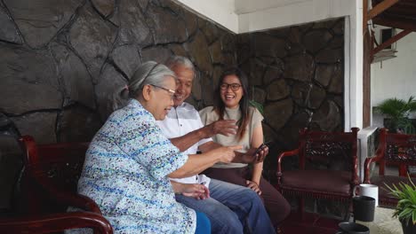 asian family with senior parents and adult daughter watching video on a mobile phone and relaxing on sofa at home together