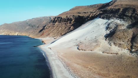 Pristine-Socotra-beach-with-white-sand,-clear-turquoise-waters,-and-a-backdrop-of-rugged-mountains,-creating-a-picturesque-coastal-landscape