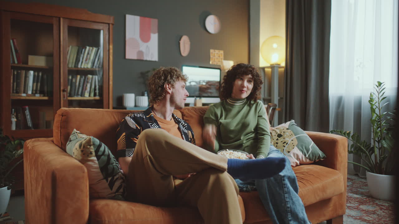 Young Couple Relaxing on Sofa, Eating Popcorn and Watching TV at Home
