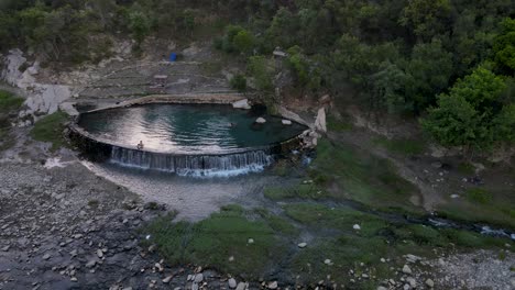 ángulo-Bajo-Aéreo-De-Aguas-Termales-Y-Puente-De-Piedra-De-Arco-Otomano,-Río-Vjose-Permet,-Albania