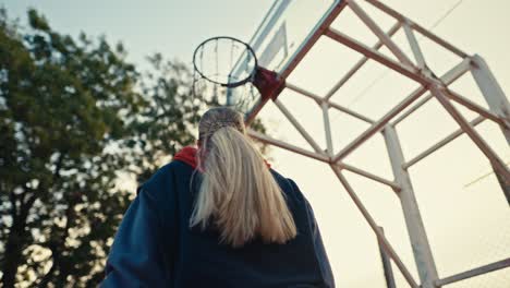 Shot-from-below-a-blonde-girl-in-a-sports-uniform-runs-with-an-orange-basketball-sword-and-throws-it-into-the-hoop-on-a-red-street-court-during-the-day