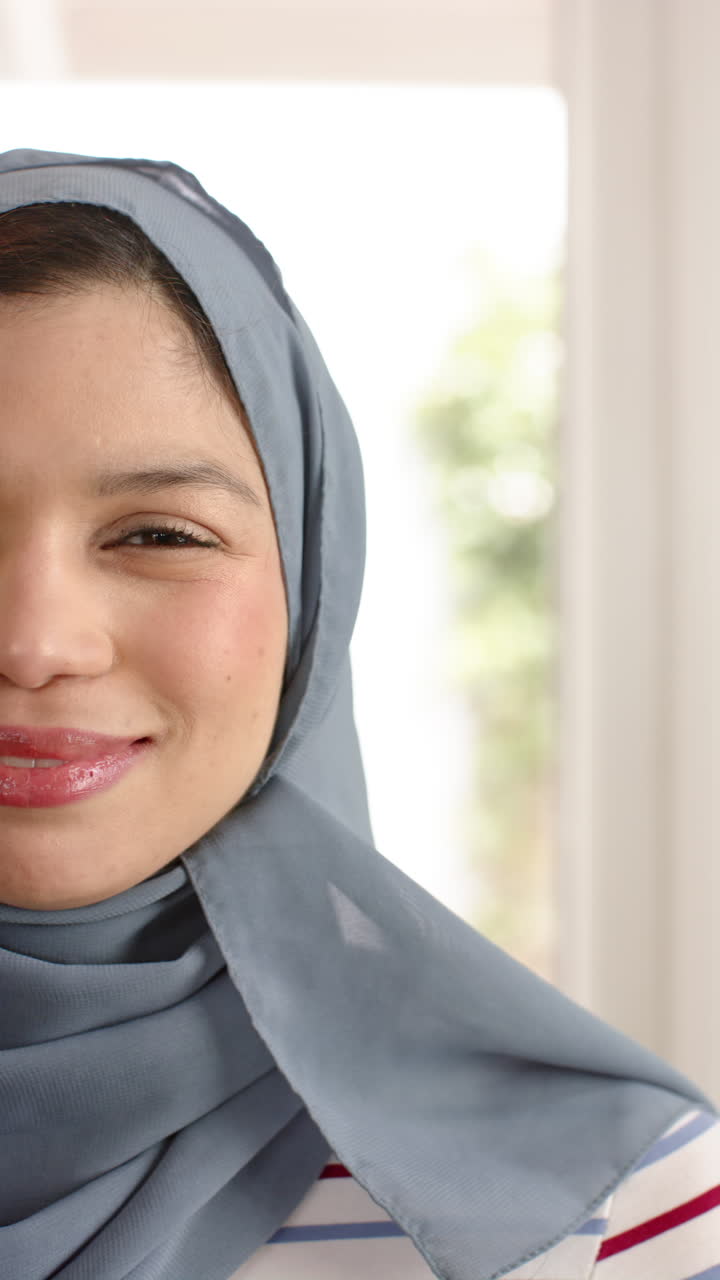 Vertical video of happy biracial woman in hijab smiling to camera at home, copy space, slow motion