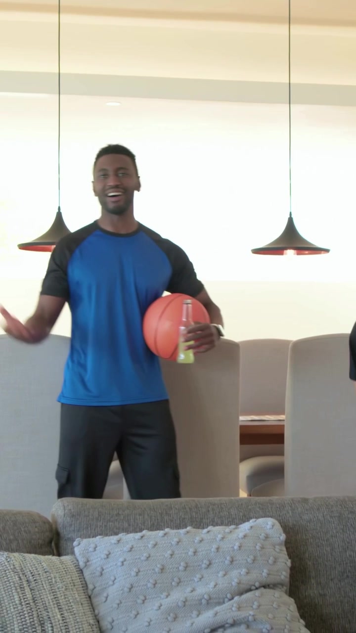 Vertical video: Grabbing snacks, blue-shirted man walking at home carrying ball, joining friend