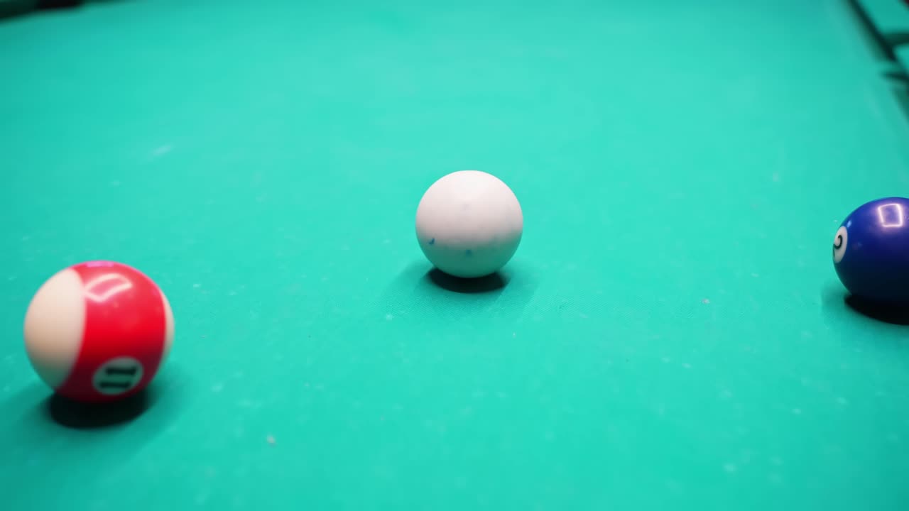 Close-up of a white cue ball resting on green pool felt with blue and red balls softly blurred at the edges, shallow depth-of-field for clean billiards b-roll