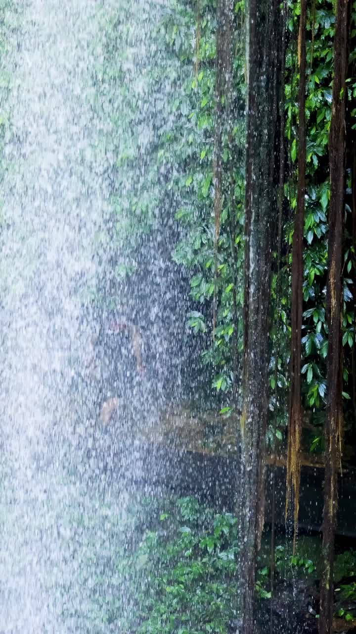 A person walks behind a waterfall in a vibrant, green forest. Natural lighting and dynamic water movement create a serene atmosphere