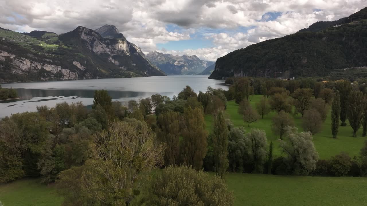 Drone slowly pulls back over lush greenery beside lake, mountains in background under cloudy sky at Walensee, Switzerland.