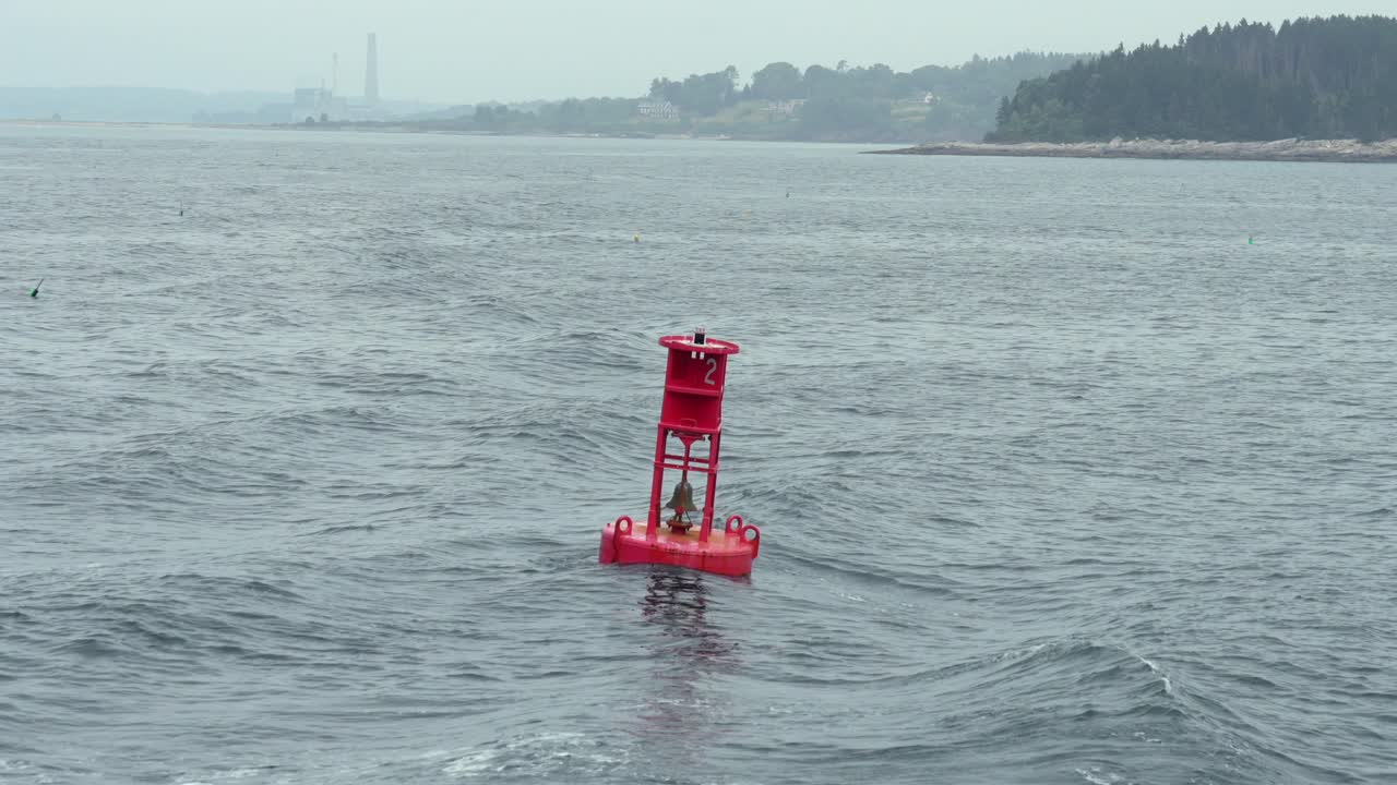 Red navigation buoy drifting on calm coastal waters with distant shoreline and light haze, capturing a quiet maritime scene with gentle motion