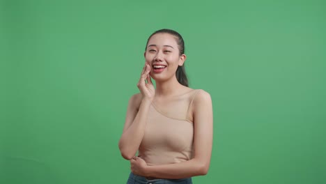 아시아 여성이 웰니스 피부 관리를 위해 파우더 얼굴을 적용하고 스튜디오에서 녹색 화면 배경에 서서 미소 짓고 있습니다.