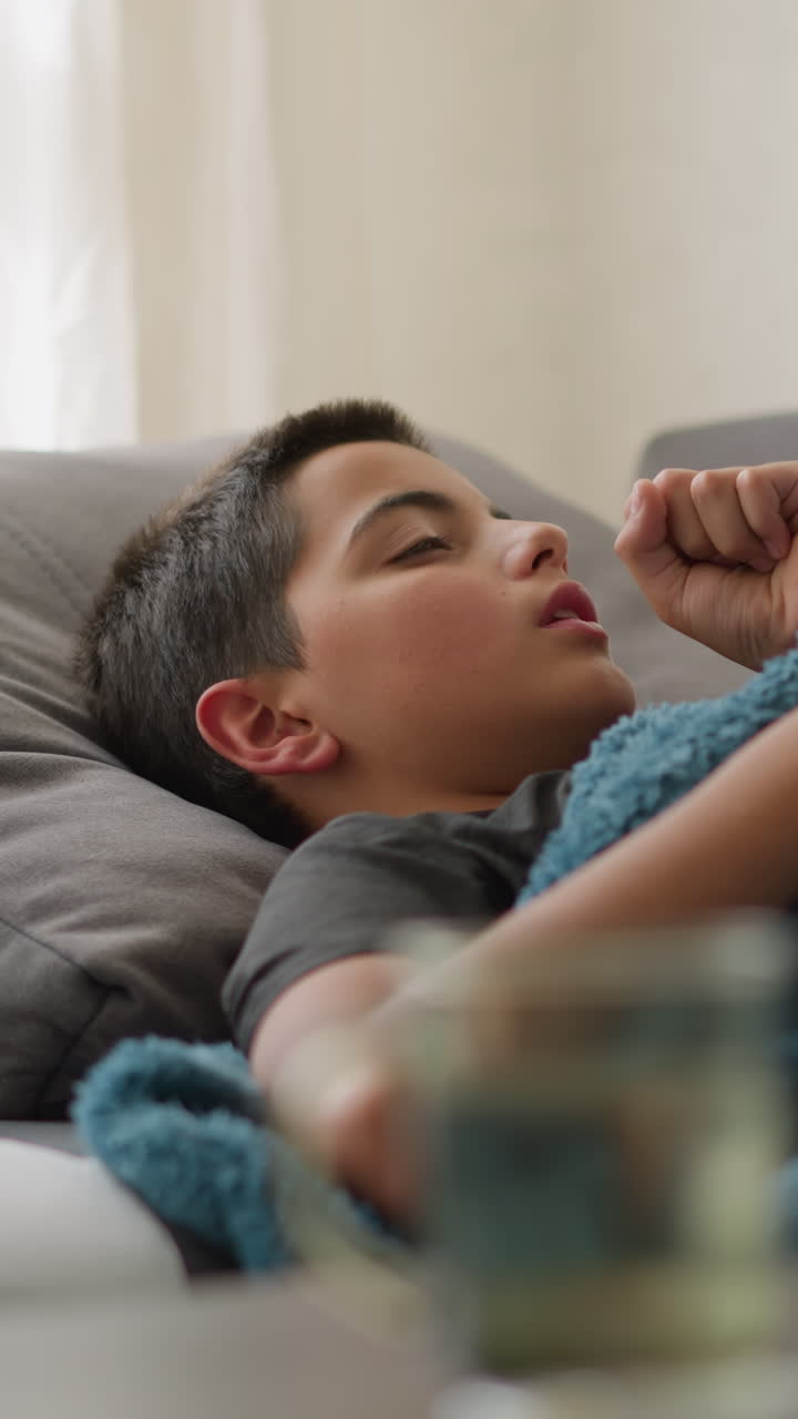 Sick kid lying on couch covered in blanket, coughing while resting hand on his head, tissue nearby and blurred view of cup and tissue on table