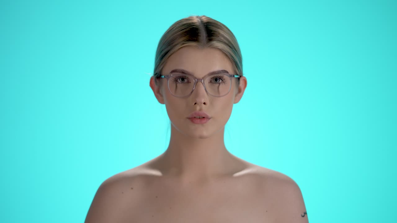 Static medium shot of a young pretty woman putting on her glasses for a clear view and looking at the camera in front of turquoise background in slow motion