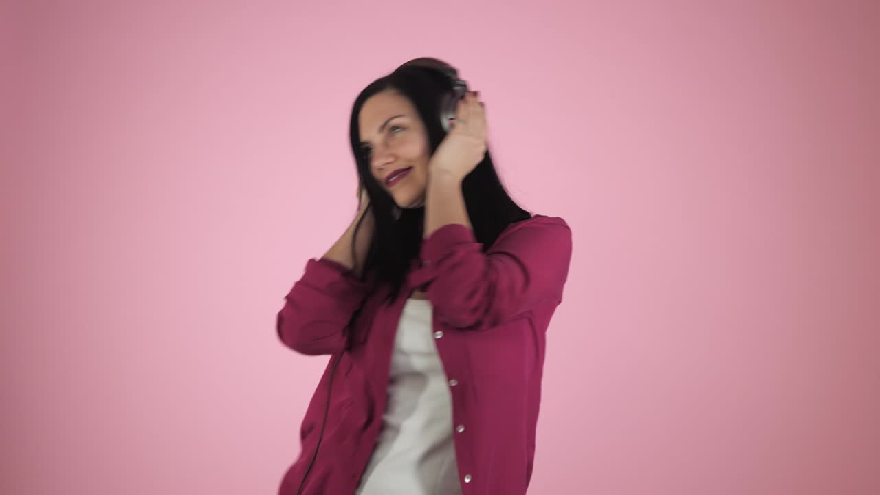 hermosa joven morena con auriculares escuchando música y cantando en fondo de color. chica feliz en camisa rosa. 4k