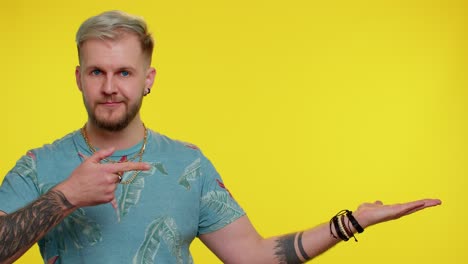 a man with blonde hair and a beard smiles and gives a thumbs up. he is wearing a blue shirt with a floral pattern and a gold chain.