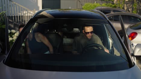 Confident-guy-in-sunglasses-with-a-beard-gets-into-the-car-with-his-girlfriend-and-fastens-his-seat-belt-before-driving