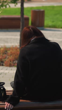 vista trasera de una mujer sentada en un parque público con su mano descansada en un banco, bolso colocado cerca, atmósfera relajada con alrededores verdes y características panorámicas del parque en el fondo