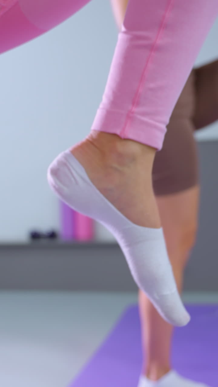 분홍색 운동복을 입은 여성이 현대적인 피트니스 스튜디오에서 친구와 함께 운동하는 클로즈업, 둘 다 균형, 힘 및 유연성에 초점을 맞추는 동기화 된 운동 루틴에 참여합니다.