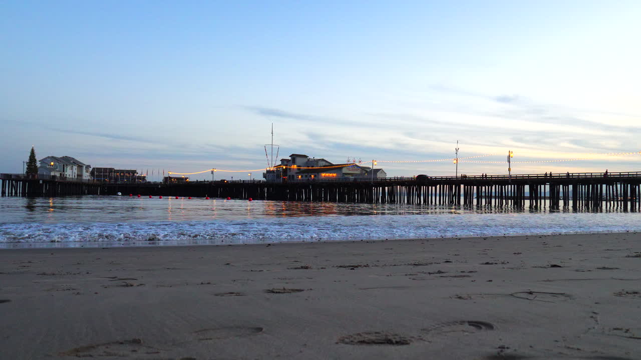 Una suggestiva spiaggia sabbiosa della California con il molo Stearns Wharf oltre le onde dell'oceano, illuminato da lanterne e luci al tramonto a Santa Barbara