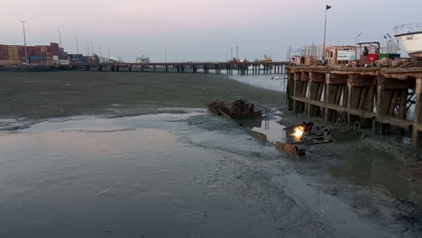 men cutting metal boat carcass with low tide in the mud, at the commercial port of guinea bissau, at dusk