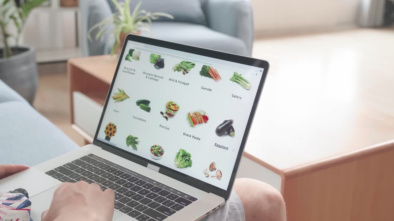 Asian Man Sitting On Couch And Using Laptop Computer Buys In Internet Shop. Male Selects Vegetables In Grocery Online Store