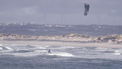 Kitesurf-En-Las-Olas-Del-Mar-Agitado-De-Peniche,-Portugal,-Molinos-De-Viento-Y-Ciudad-En-Segundo-Plano