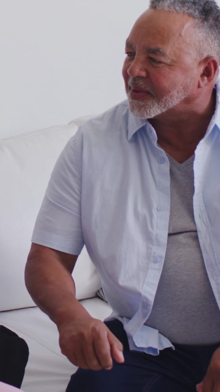 Video verticale: Donna che si appoggia e parla, uomo che si sposta e si siede sul divano in camicia blu unendosi alla conversazione.