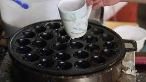 kanom krok thai coconut pudding pancakes, pouring a cup of white coconut milk on hot traditional krok pan