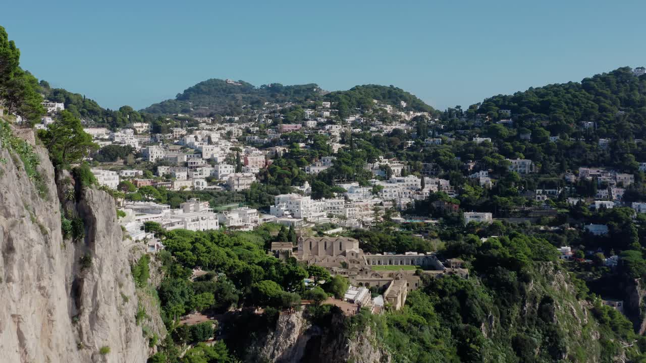 Volando detrás de un escarpado acantilado de piedra caliza y revelando un hermoso pueblo en la isla de Capri. Pueblo italiano en la isla con edificios de tonos blancos.