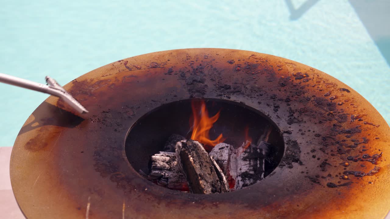 Primo piano di una piastra di cottura con braciere, con un uomo che raschia i residui di cibo, piscina sullo sfondo, fiamme al centro del barbecue