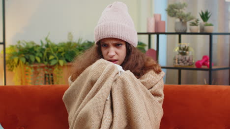 sick child girl kid wear hat wrapped in plaid sit alone shivering from cold on sofa drinking hot tea