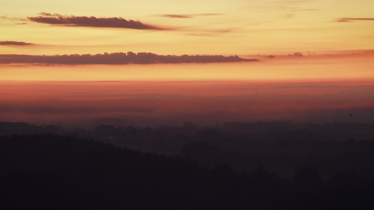 Timelapse of a sunset over a misty landscape. Layers of orange, pink and purple hues blending into the horizon. Dark silhouettes of hills in the foreground.
Fog rolling over valleys during twilight.