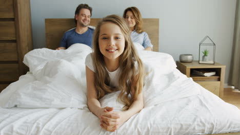 cute little girl lying and smiling at camera, her parents are lying under the blanket on the bed behind her