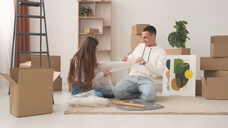 Zoom-In-Shot-Of-Young-Couple-In-A-New-House-Sitting-On-The-Floor-Unrolling-Bubble-Wrap