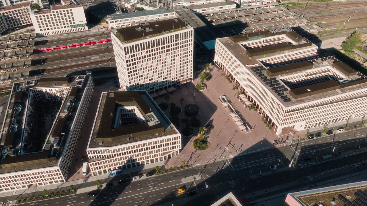 Drone pulls back above the modern station plaza, revealing geometric office blocks, open square and passing red train in the rail district