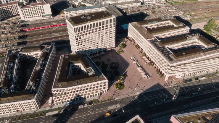 Un drone si allontana dalla moderna piazza della stazione, rivelando blocchi di uffici geometrici, una piazza aperta e un treno rosso di passaggio nel quartiere ferroviario