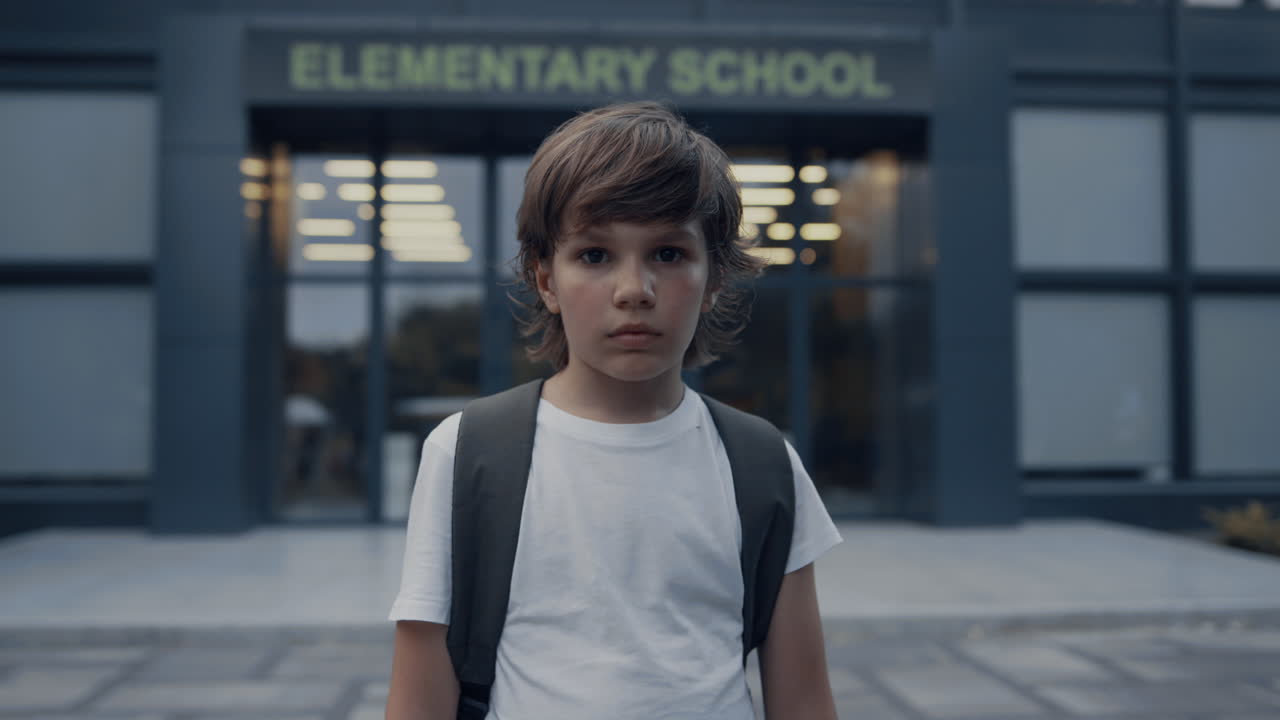 Sad elementary age boy near modern school entrance. Teen student looking camera.