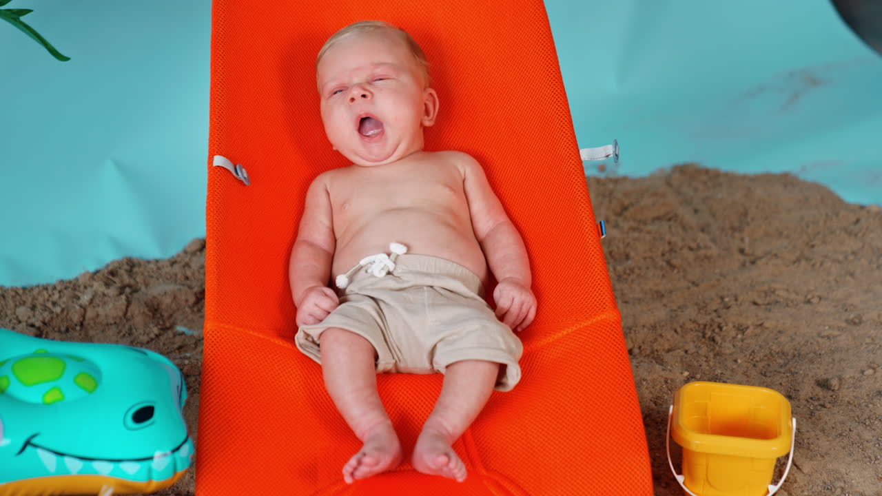 Relaxed yawning baby boy lying in the orange chair. Sand and swimming circle around the kid.