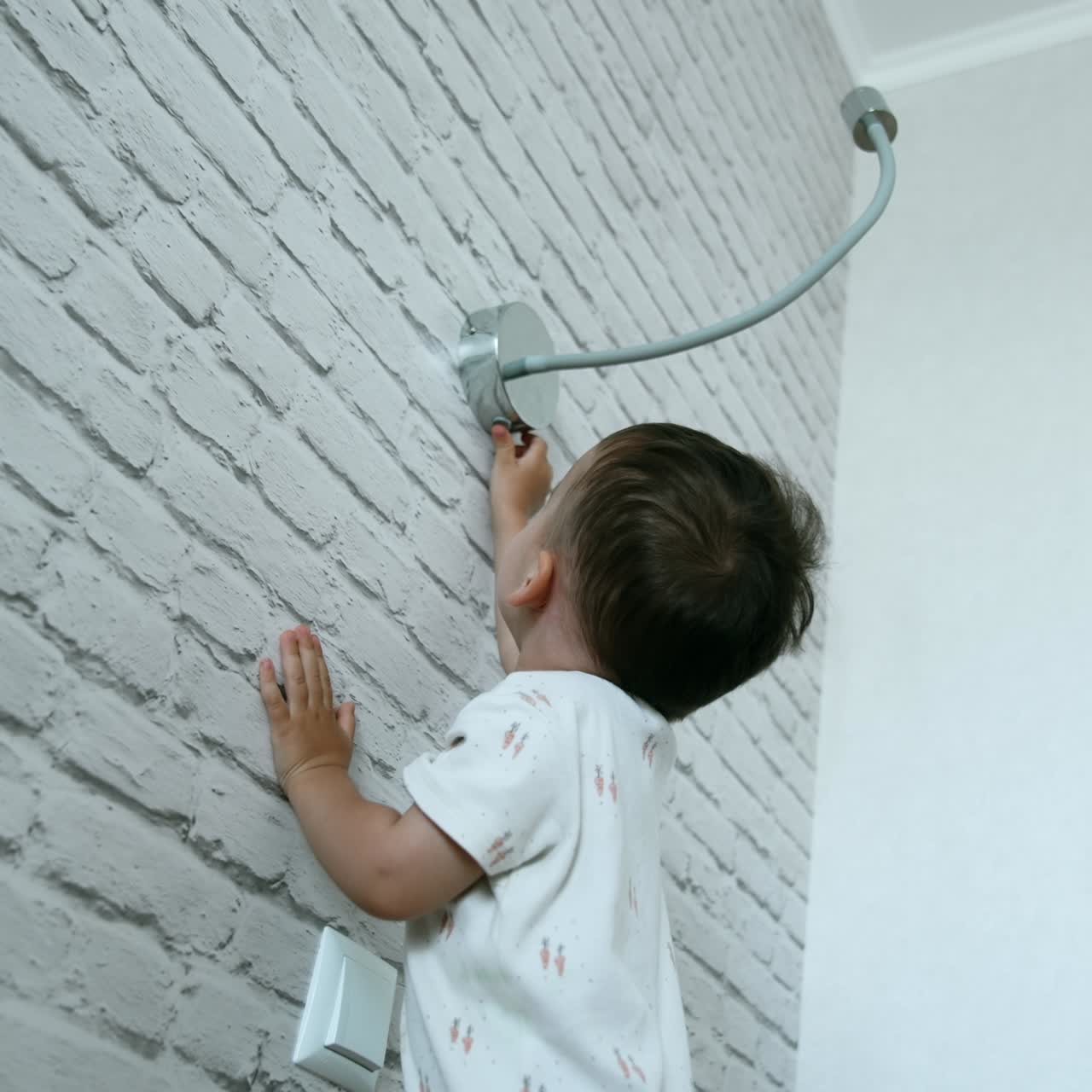 Little cute boy stands holding by the white brick wall. Curious toddler reaches the lamp above him to switch it off and on again. Low angle view