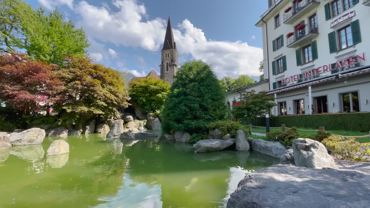Toma estática, día soleado parcialmente nublado, estanque de carpas koi en un jardín japonés con campanario de iglesia y Hotel Interlaken, atmósfera suiza serena.