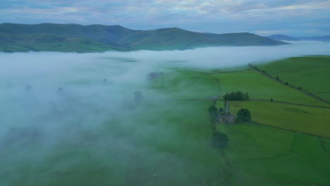 flying over green farmland with slow pan revealing mountain range and dense fog bank at sunrise