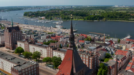 Aerial-view-of-city-lying-on-bank-of-river.-Main-wide-street-with-massive-buildings.-Fly-above-Saint-Marys-church-red-tiled-roof-and-tower